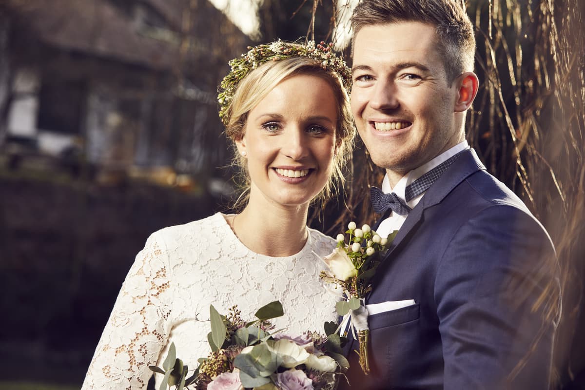 Strahlendes Brautpaar im Porträt: Braut mit Blumenkranz und Spitzenkleid, Bräutigam mit Fliege und Blumenanstecker