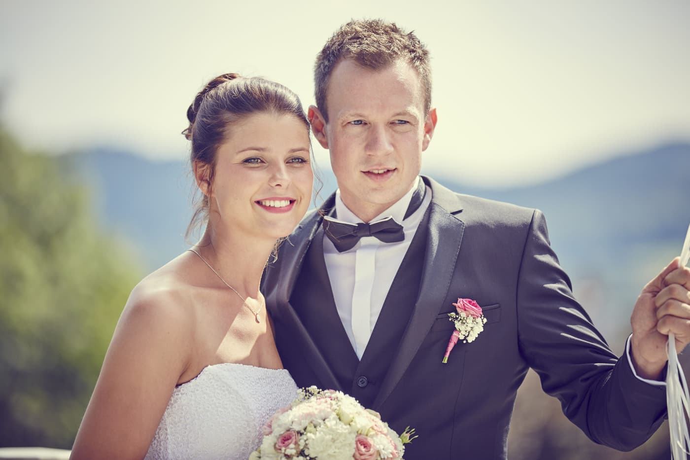 Brautpaar im sommerlichen Porträt vor Hügellandschaft, Bräutigam mit Fliege und rosa Rosenanstecker, Braut mit Brautstrauß