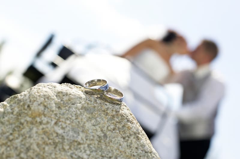 Zwei gravierte Eheringe auf einem Stein, dahinter unscharf das Brautpaar beim Kuss – Detailaufnahme am Hochzeitstag